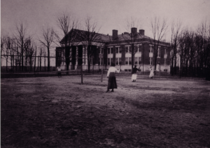In black and white, four women are playing tennis in front of a building, with several bare trees surrounding the area.